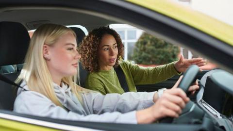 A learner driver with an instructor who is pointing and fiving instructions while the driver holds the steering wheel and looks ahead in front.