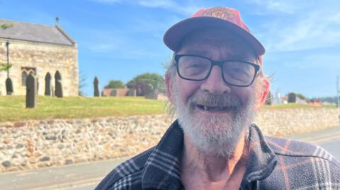 An elderly man has a white beard and wears black-rimmed glasses, a Manchester United cap and a dark, checked shirt. He is standing beside a road under a blue sky. In the background, gravestones stand on green grass behind an old stone wall and part of a stone village church can be seen.
