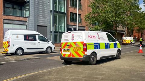 three police vans on a city street, with an officer behind a police tape across the road