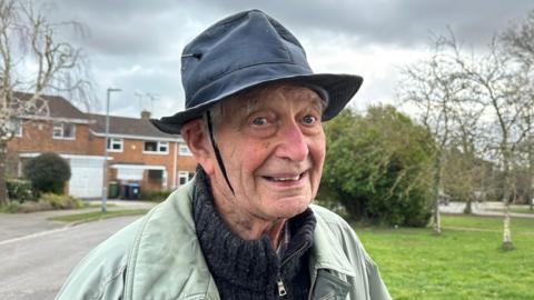 91-year old Glyn Parry is stood outside smiling at the camera. He is wearing a hat with a piece of shoelace tied underneath his chin to keep his hat on.