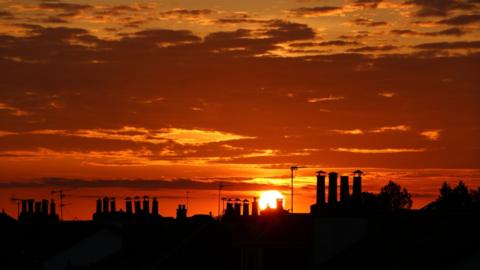A golden sunset with silhouetted rooftops and chimneys in the foreground