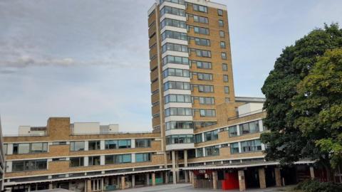 A high rise accommodation block overlooks the university's main square with a large tree at the end.