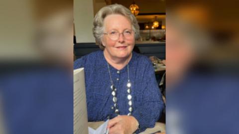 Joan Bell has short grey hair and glasses and is wearing a blue jumper. She is smiling in the photo.