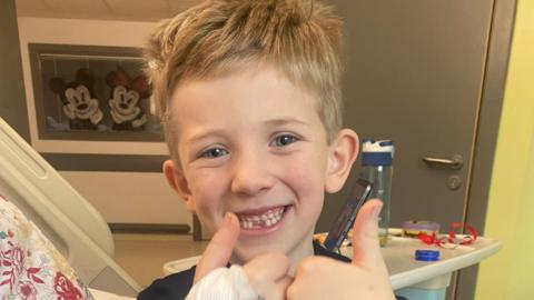 Lewis Danton sitting in a hospital bed in his room. He is smiling at the camera and is holding his thumbs up. 
