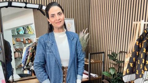 A woman with long dark hair is wearing a white top, brown skirt with white spots and a blue denim jacket. She is standing with a mirror behind her, wooden walls and plants.