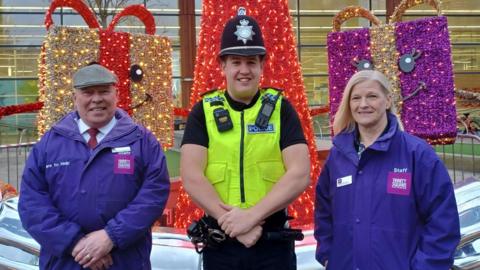 A police officer in full uniform stands next to a man and woman who are wearing purple coats with the logo 'Trinity Square'.