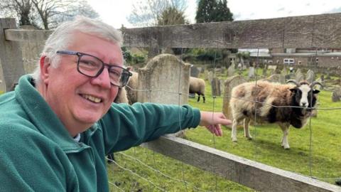 David Edwards is smiling and leaning on a fence next to one of the sheep. He is wearing glasses and a green fleece. Behind the mesh fence there is a brown woolly sheep with a black and white face and horns standing on grass. Behind it there are several headstones. 