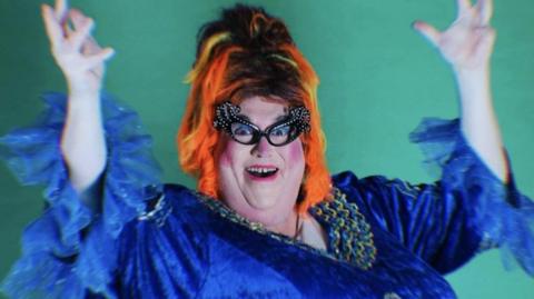A pantomime dame, in the style of Dame Edna Everage, wearing a blue dress with bright orange hair and large glasses.