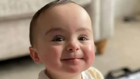 Close up of the smiling face of the late nine-month-old Genevieve Meehan. She has brown eyes and is wearing a lemon and white outfit.