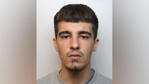 Police mugshot of Kurtis Knee with a shaved moustache and goatee beard, dark hair and dark eyes, wearing a grey top