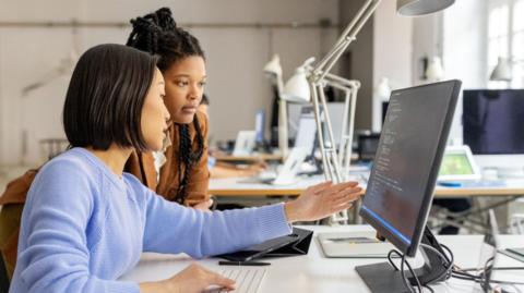 Two women sit at computer with desks in the background.