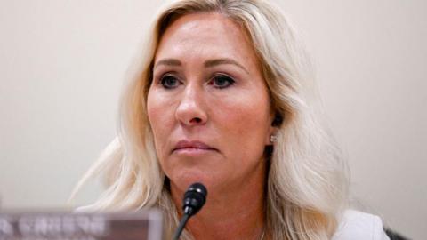 Marjorie Taylor Greene sits at a committee hearing table, with a microphone and name placard placed before her.