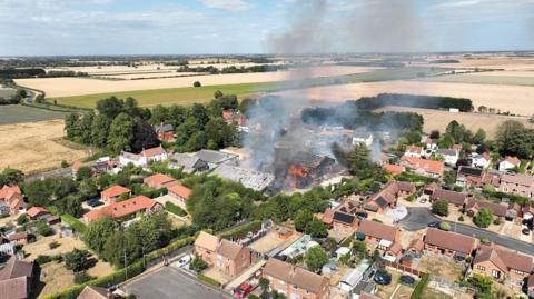 A drone shot of a large fire in buildings in the middle of a village. The flames are clearly visible.