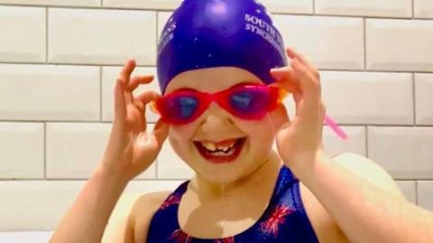 A young girl grinning in swimming costume, goggles and hat.