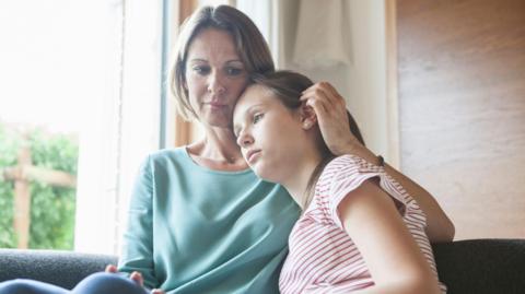 A mother and daughter are sat on a sofa looking downcast. The mother is wearing a blue shirt and has short hair. The daughter is wearing a red and white striped T-shirt.