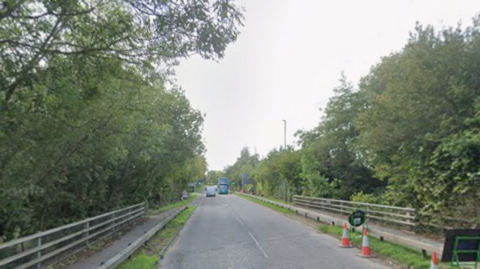 A large road with railings and trees on either side. Some traffic cones can be seen to the right of the carriageway.