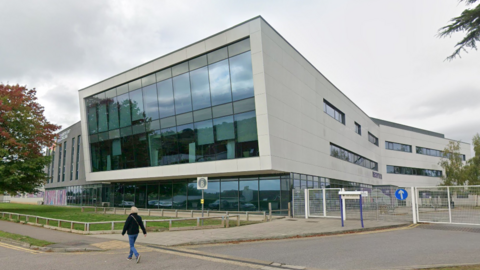A college building with a large glass front and grey walls. A person is walking in front of the building.