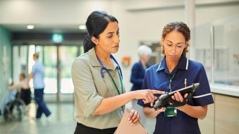 Staff nurse consulting ward doctor - both are women. One is standing holding a tablet, whilst another is standing next to her pointing at the tablet.