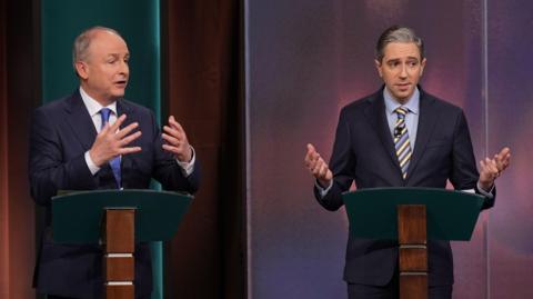 Micheál Martin and Simon Harris at the podium during a TV debate. Both men  are wearing dark suits and expressively using their hands 