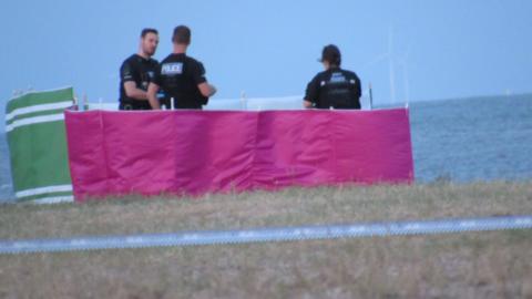 Three police officers standing on the beach in West Mersea. They are surrounded by green, pink and white windbreaks. Blue and white tape - indicating a cordon is in place - is pictured in the foreground.