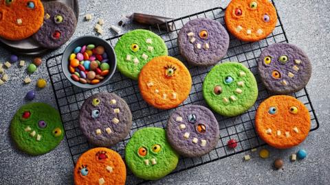 A cooling rack with several orange, purple and green cookies with eyes and teeth.