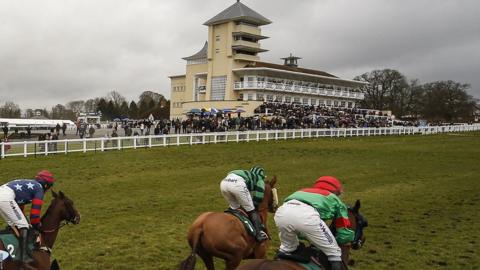 Horseracing at Towcester Racecourse. Three horses are visible - two with jockeys in green colours almost neck-and-neck and a third about a length behind with a jockey in blue colours with white stars. The cream-coloured pavilion with a four-storey tower is visible behind.