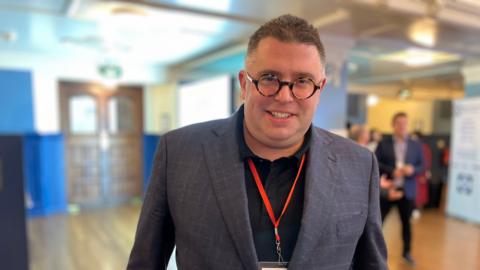 Former fraudster Alex Wood is pictured at the Oxford Fraud Conference. He's wearing an orange lanyard, grey checked suit and round tortoiseshell glasses.