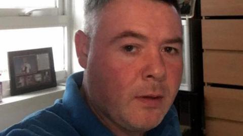 A head and shoulders close up of a clean shaven man in a blue polo shirt looking serious