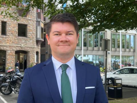 Deputy Sam Mézec is pictured wearing a green tie and a navy blue suit with a light blue shirt. He has short dark hair and he is smiling. He is standing outside the government of Jersey building.