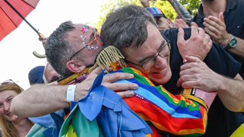 A person wearing a jacket in the colours of the pride flag hugs Budapest Mayor Gergely Karacsony, who is wearing a black T-shirt and glasses.