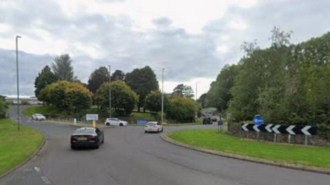 Stainburn roundabout in Workington. There is a green patch of grass in front of the roundabout. There are a few cars travelling on the roundabout. There are trees in the background.
