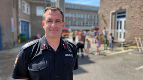 The image shows Paul Brown standing in the foreground wearing a dark fire and rescue service uniform, facing the camera in an outdoor courtyard or service area between two stone buildings. There are children behind him near a fire engine.