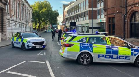 Police cars parked in the middle of a city centre street. It is daytime. Police officers are in the background.