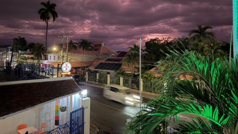 A view of a street with a purple sky, buildings and a car moving along a road