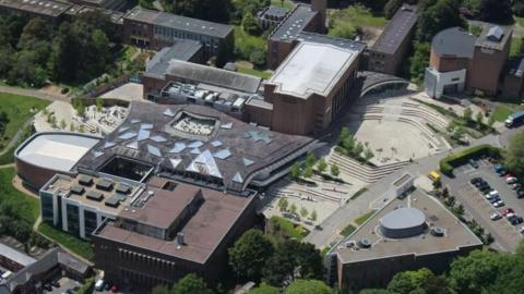 An aerial view of the Univercity of Exeter campus, showing various buildings, roads, public spaces and trees.  