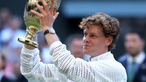 A young man hold a golden trophy up in the air. He is wearing a netted jacket and is smiling.