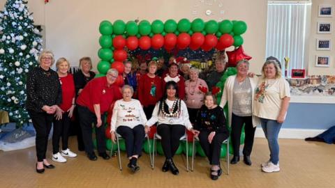 Nearly 20 people are lined in front of green and red balloons arranged in a square at a gathering of a group at Parkside Community Centre . There is a tall Christmas tree at the left corner of the room. People are smiling. Denise Cameron is the last one standing one the right, in white T-shirt, blue jeans and white trainers. Her hair reaches her shoulders and is wavy and blonde. 