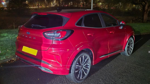 A red hatchback car is parked by the side of a road. The rear number plate has been blanked out.