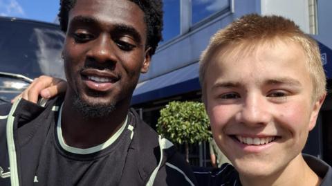Norwich striker Mo Touré (left) is wearing a black T-shirt. He is standing next to Ranulph Bacon, who is wearing a navy polo shirt. The pair are smiling at the camera.