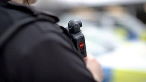 A close up of a police officer's shoulder. They are wearing a bullet proof vest with a radio mounted on it. A police car is blurred in the background.