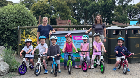 A row of children about age four and five on bikes, two women standing behind, at a playground. Trees and blue skies seen behind.