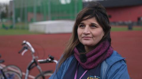 An Asian woman wearing a scarf and navy jacket, looks to the left of the camera as she sits on a bike on a running track