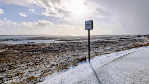A passing place on an empty road through an area of moorland and lochs in Shetland. The landscape has a dusting of snow.