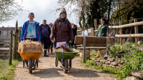 A man and woman walking with wheelbarrows in farm with people in the background.