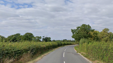 A country lane snakes to the right with trees and hedges on the right hand side and a hedge along a field is on the left. Other trees can be seen in the distance on the left hand side of the image.