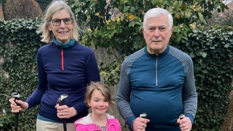 Two grandparents, a man and a woman, wearing walking gear and holding walking poles. Their granddaughter, wearing a pink coat, is stood in between them.