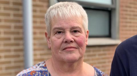 A woman with short white hair and brown eyes looks into the camera. Cut off at the shoulder you can see she wears a blue, orange and white coloured top. A mans shoulder can be seen to her left, he wears a navy top. Behind her, out-of-focus is a red bricked wall. A partially opened translucent window is positioned over her left shoulder. 