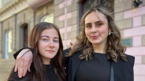 Summer and Maya are standing close together outdoors in front of a stone building. Maya has an arm around Summer's shoulder. Both are wearing dark clothing, and the background shows a paved area and architectural details including arched windows and stone walls.