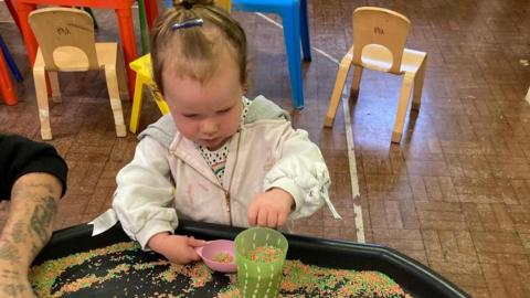 A baby girl wearing a grey top pours orange and green coloured rice granules into a green cup 