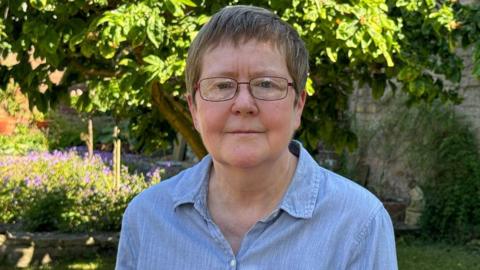 A woman with dark hair and glasses is looking at the camera. She is wearing a blue top. She is standing in a garden with a stone wall, surrounded by trees and flowering plants in the background.